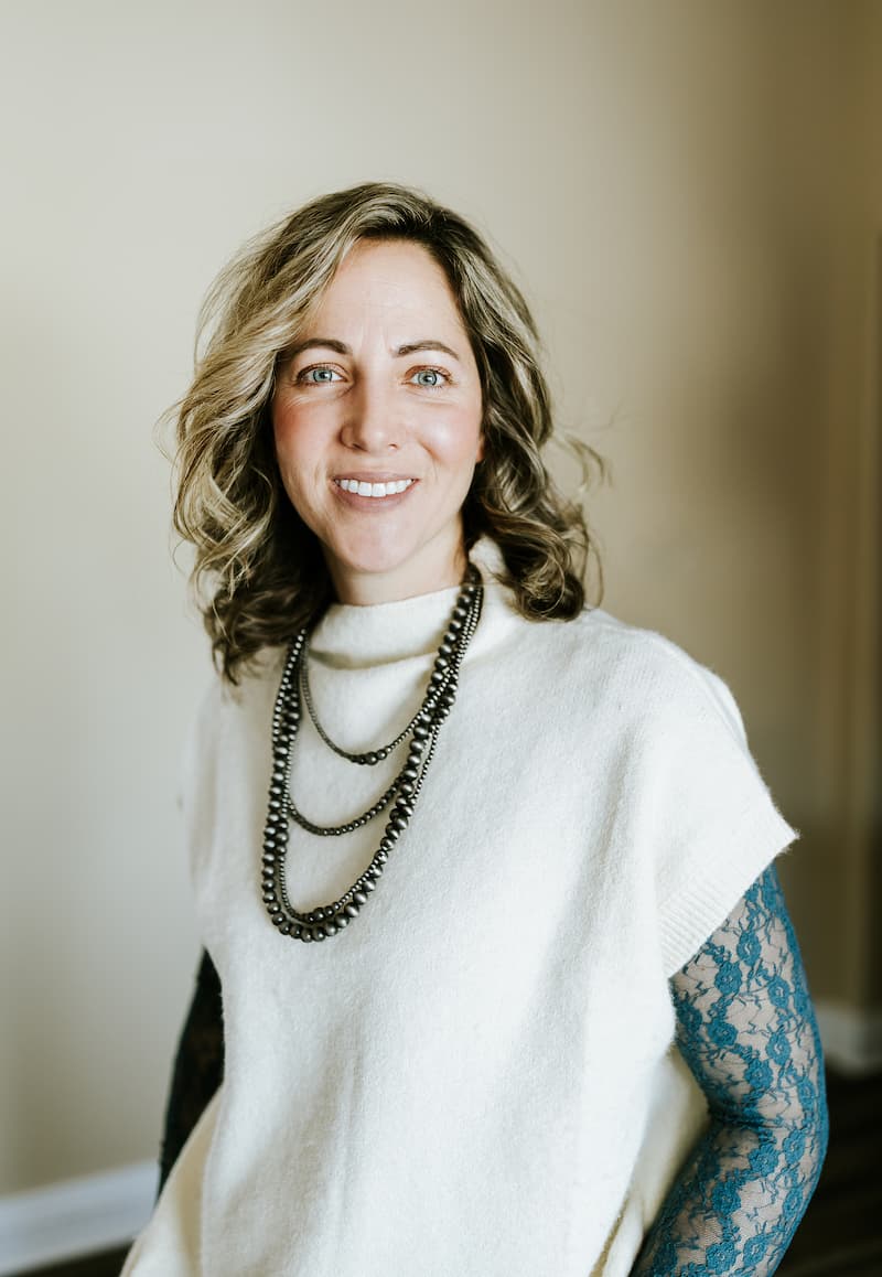 Woman with wavy blonde hair wearing a white top, layered necklaces, and blue lace sleeves, smiling in a neutral indoor setting.
