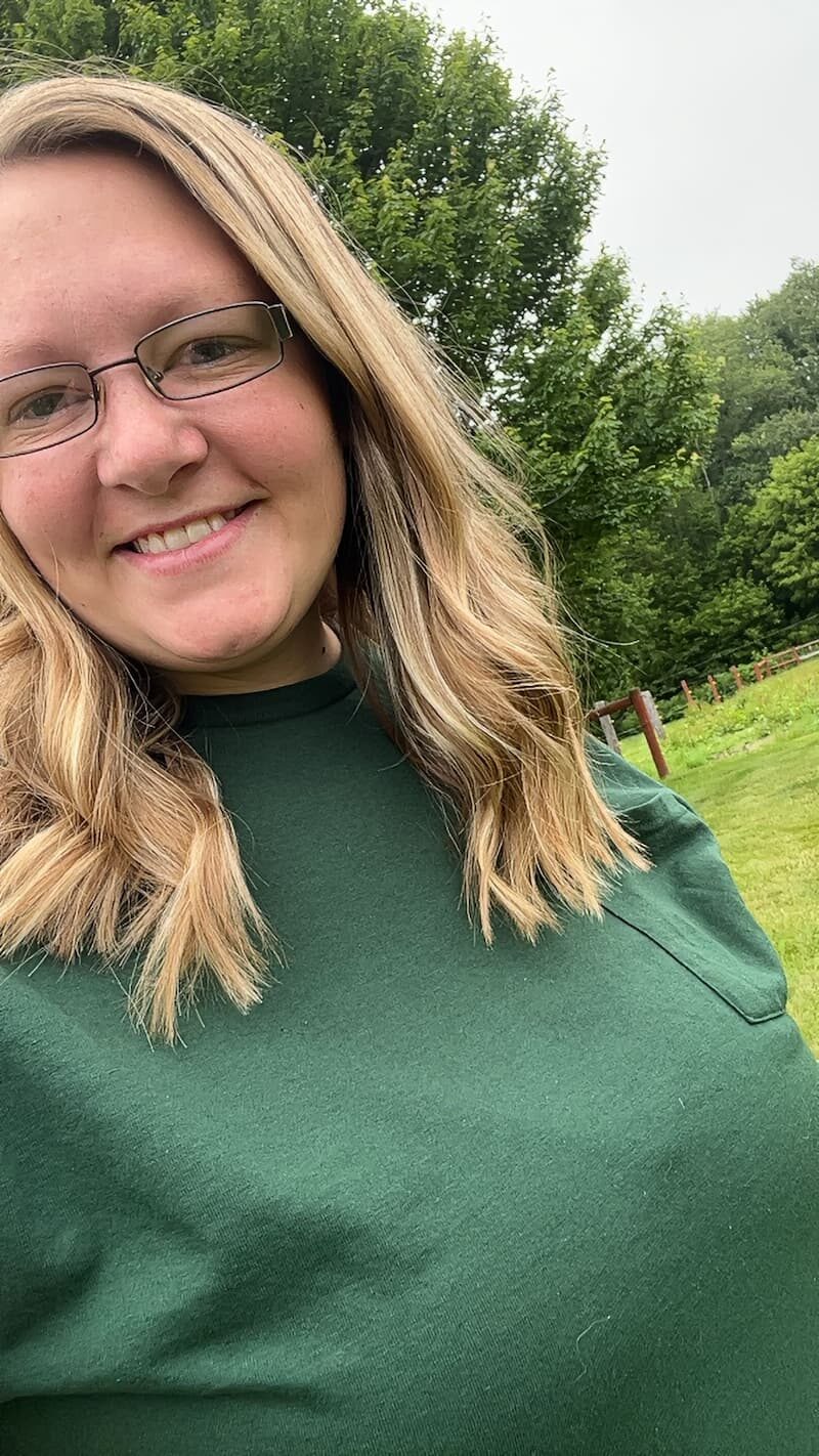 Woman with blonde hair and glasses wearing a green shirt, smiling outdoors with trees and grass in the background.